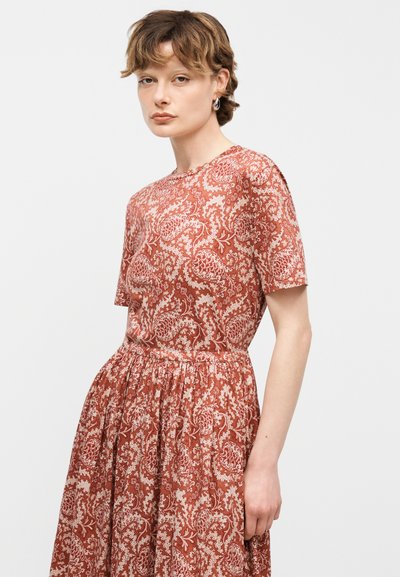 Νεαρή γυναίκα με κοντά μαλλιά, φοράει φόρεμα με κοντά μανίκια σε σκουρόχρωμο πορτοκαλί με φλοράλ μοτίβο, στέκεται μπροστά σε απλό φωτεινό φόντο.