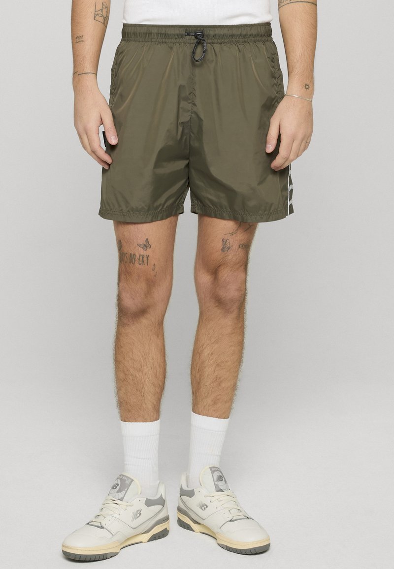 Mann trägt olivgrüne Shorts mit Kordelzug, weiße Socken und weiße Turnschuhe, steht vor einfachem Hintergrund.