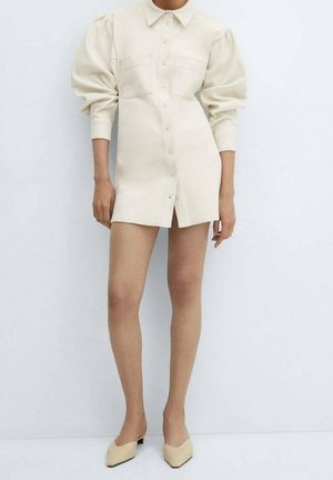 Crèmekleurige jurk met knopen, pofmouwen, twee borstzakken en een rechte snit. Gecombineerd met nude schoenen, tegen een neutrale achtergrond.