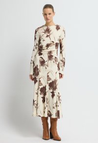 Kwiatowa sukienka z długim rękawem o kremowym tle i brązowym abstrakcyjnym wzorze kwiatowym, z dopasowaną talią i zwiewną spódnicą. Brązowe botki do kolan.