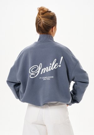 Personne avec les cheveux attachés en chignon, portant un sweat-shirt gris avec un texte blanc disant "Smile! It looks good on you" et un pantalon blanc, vue de dos.