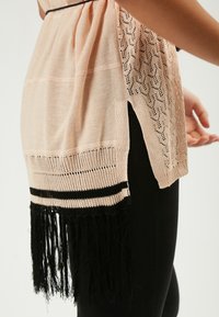 Hellrosa gestrickter Schal mit offenem Spitzenmuster, schwarzem Streifen am Saum und fransigen Kanten. Über schwarzen Hosen getragen, wobei die Seitenansicht zur Schau gestellt wird.