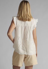 Femme aux cheveux blonds mi-longs portant un haut sans manches brodé blanc et un short beige, se tenant devant un fond gris.