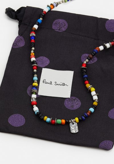 Fargerik perlekjede med ulike materialer og former, med en sølvcharms, plassert på en svart prikkete stoffposen merket "Paul Smith."