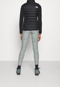 Svart pufferväst med horisontell quiltad design, grå texturerade leggings och blå sportskor. Synlig logotyp på västen.