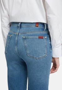 Powblakłe niebieskie dżinsy z denimu mają pięć kieszeni, wysoki stan oraz małą czerwoną metkę z cyfrą "7" na tylnej taśmie w pasie.