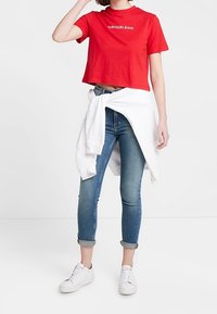 Rød crop-top med "Calvin Klein Jeans" logo, parret med lyseblå skinny jeans og en hvid sweatshirt bundet om taljen. Hvide sneakers.