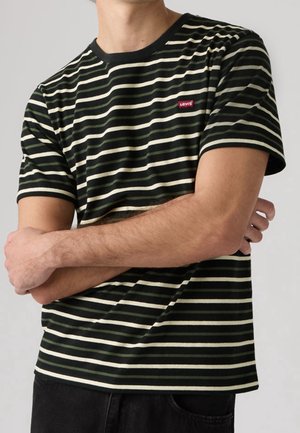 Person iført en sort- og hvidstribet Levi's t-shirt med armene krydset, synlig fra skuldrene til taljen.