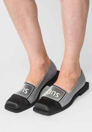 Coppia di scarpe slip-on grigie e nere con punta rigata e pannelli in maglia bianca che mostrano la parola "SUE", indossate a piedi nudi.