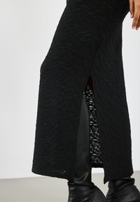 Svart strikket kjole med et teksturert m�ønster, med en sideåpning. Stoffet har et lett, mykt utseende, supplert av tettsittende svarte støvler.