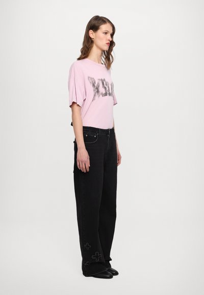 Ljusrosa oversize t-shirt med en svart grafisk design, matchad med svarta vida jeans med broderade detaljer och en slät textur.