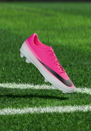 Adidași de fotbal roz aprins cu swoosh negru, plutind deasupra unui câmp de iarbă verde lângă marcajele albe de pe teren.