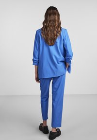 Completo blu composto da blazer a maniche lunghe con vestibilità rilassata e pantaloni affusolati abbinati, realizzato in tessuto liscio, indossato con scarpe nere.