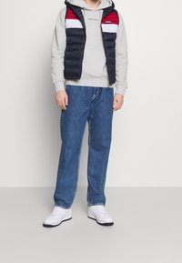 Padded navy vest with red and white accents over a gray hoodie, paired with loose-fitting blue jeans and white sneakers with black soles.