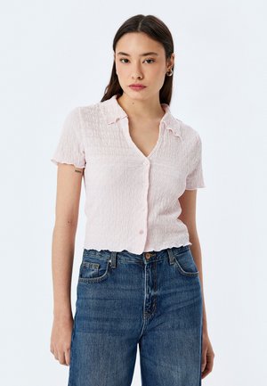 Blouse à manches courtes, avec col, de couleur rose clair et à motif texturé, équipée d'un devant à boutons et d'un ourlet festonné, associée à un jean bleu.