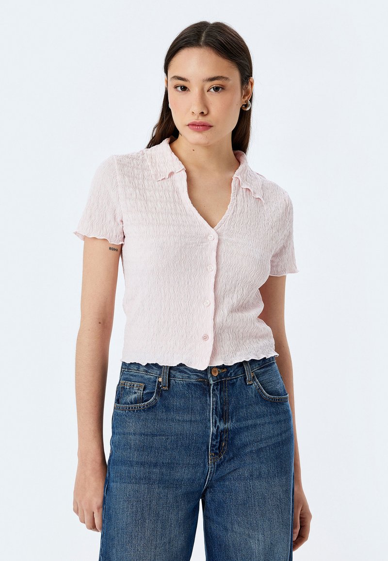 Blusa a maniche corte con colletto di colore rosa chiaro, caratterizzata da un motivo testurizzato, con chiusura a bottoni frontale e orlo scallop, abbinata a jeans blu.