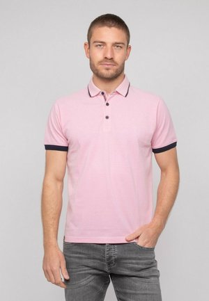 Homme portant un polo à manches courtes rose clair avec un bord foncé au col et aux manches, debout avec une main dans la poche devant un fond uni.