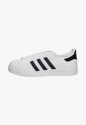 Witte Adidas sneaker met zwarte strepen, rubberen neuzenkap en een bovenwerk van gestructureerde stof, weergegeven vanaf de zijkant op een witte achtergrond.