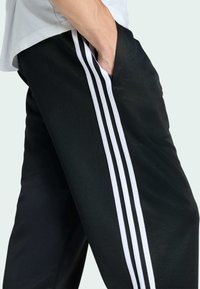 Black athletic pants featuring white three-stripe design along the sides; made of smooth fabric with side pockets and elastic waistband.