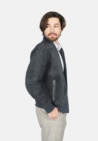 Šedá suede bomber bunda s ribbovaným límcem, zipovými kapsami a měkkou texturou, nošená přes košilí s límcem a kostkované kalhoty.