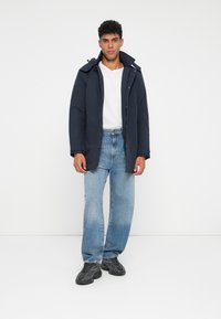 Giubbotto imbottito blu navy con zip frontale, collo ampio e tasche, abbinato a jeans larghi in denim chiaro e sneakers nere texturizzate.