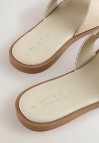 Šviesiai smėlio spalvos šlepetės su zomšiniu viršumi ir tekstūruotu padu. Padas rudas ir turi iškeltą prekės ženklo tekstą "NEXT FOREVER COMFORTS."