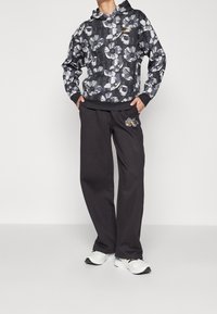 Person, der einen schwarzen Hoodie mit großen weißen Blumen und schwarze Hosen mit floralem Logo trägt, steht mit den Händen in den Taschen und weißen Sneakers.