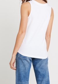 Débardeur en coton blanc avec un col rond. Le dos présente une texture lisse et un ourlet légèrement courbé. Associé à un jean en denim bleu clair.