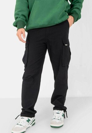 Black cargo pants with multiple side pockets, tapered leg design, and adjustable cuffs; worn with a green hoodie and white-green sneakers.