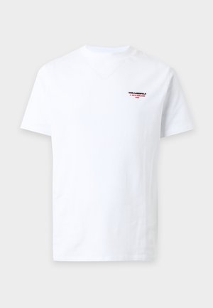 Jeune homme debout les mains dans les poches, portant un t-shirt blanc à manches courtes, un pantalon noir et des baskets noires et blanches.