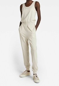 Mörkhyad person som bär en beige ärmlös topp, matchande högmidjade joggarbyxor och beige snörskor, står med händerna i fickorna.