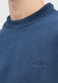Záber na krk a rameno, ktoré nosí modrý svetrový triko s okrúhlym výstrihom, na hrudi je malá vyšívaná šedá logo ellesse.