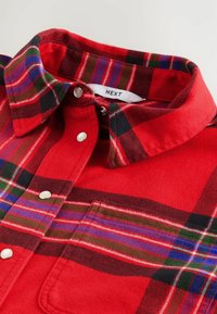 Rød ternet flannel-skjorte med knapkrave. Har flerfarvede mønstre med blå, grønne og sorte accenter. Rundet kant, sølvknapper.
