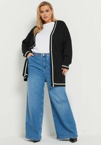 Svart ribbad cardigan med vit kant, bärs över en vit skjorta, ihop med vida blå jeans. Texturerat tyg och avslappnad passform.