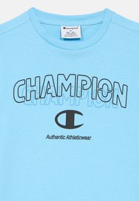 Ανοιχτό μπλε βαμβακερό μπλουζάκι με την επιγραφή "CHAMPION" σε έντονα μαύρα γράμματα και μαύρο λογότυπο, με την ένδειξη "Authentic Athleticwear" τυπωμένη από κάτω.