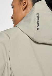 Gros plan sur une personne portant une veste à capuche beige clair en tissu texturé avec le logo "Icepeak" sur la capuche.
