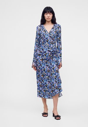 Femme aux longs cheveux noirs portant une robe midi à manches longues bleue à fleurs et des sandales noires à talons, debout devant un fond uni.