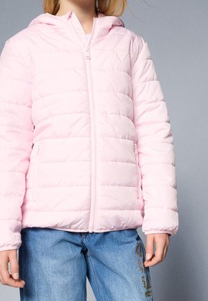 Lapsi, joka pukeutunut vaaleanpunaiseen tikattuun takkiin, jossa edessä vetoketju, ja sinisiin farkkuihin, joissa toisessa lahkeessa koristeellinen kirjainpainatus.