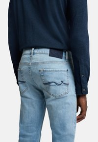 Jeans in denim azzurro con design di onde ricamato sulle tasche posteriori e un'etichetta scura. Indossati con una camicia a maniche lunghe blu navy.