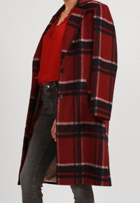 Cappotto di lana a quadri rossi e neri con una vestibilità sagomata, caratterizzato da un revers a incavo e due bottoni, indossato sopra una maglia rossa e pantaloni grigi.