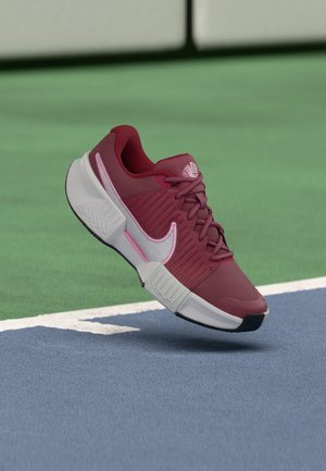 Bordowy sneaker z białą podeszwą i różowymi akcentami unoszący się nad niebiesko-zieloną linią graniczną kortu tenisowego.