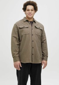 Camisa de franela marrón con dos bolsillos en el pecho, botones negros y mangas largas. Llevada sobre una camiseta oscura, combinada con pantalones negros.