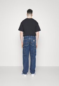 Pantalones cargo de mezclilla azul con múltiples bolsillos, corte holgado y puños ajustados, combinados con una camiseta negra de manga corta y zapatos blancos.