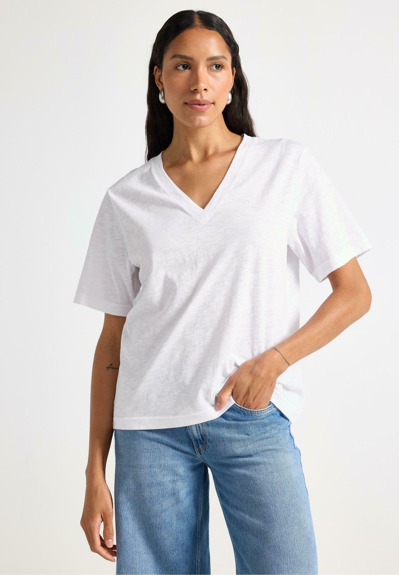 T-shirt bianco a maniche corte con scollo a V realizzato in tessuto leggero, abbinato a jeans a gamba larga blu chiaro. Il modello è in piedi con le mani in tasca.