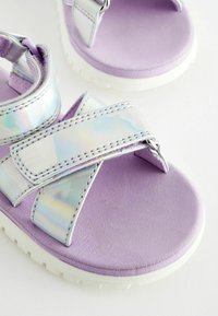 Lila sandal med iriserande remmar, med en dämpad sula och vit gummiyttersula. Remmarna har sömnadsdetaljer för stöd.
