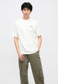 Hvid bomulds T-shirt med korte ærmer, der har rødt trykt tekst på venstre bryst. Brugt sammen med olivengrønne cargobukser med flere lommer.