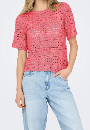 Femme portant un haut court à manches courtes en crochet rose corail et un jean taille haute bleu clair, une main dans la poche, sur fond uni.
