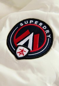 Vit tyg med en cirkulär broderad logotyp. Mörk marinblå kant med texten "SUPERDRY" längst upp och röda accenter som formar ett stiliserat bokstav "A."