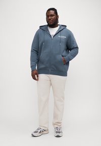 Blue zip-up hoodie with a front pocket, drawstring hood, and printed logo. Paired with cream-colored pants and gray sneakers.