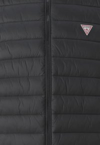 Vesta puffer neagră cu un design matlasat, având un fermoar frontal și un mic logo roșu în formă de triunghi pe partea stângă. Textură netedă și lucioasă.